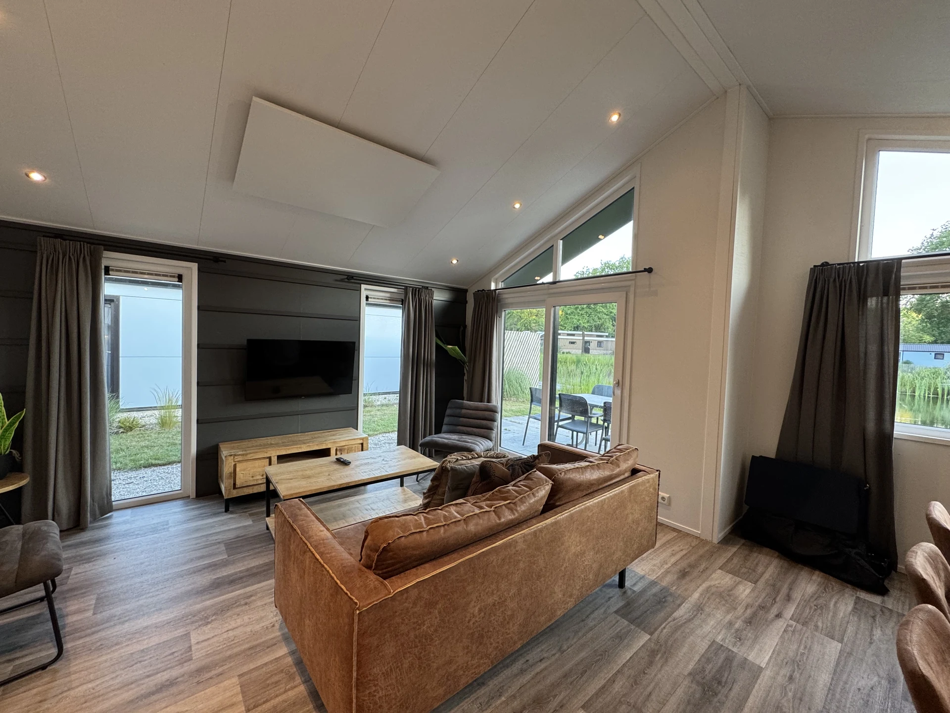 Wohnbereich eines Chalets im EuroParcs Kaatsheuvel mit Gewölbedecke, Cognac-Ledersofa, Wand-TV und großer Schiebetür zur Terrasse