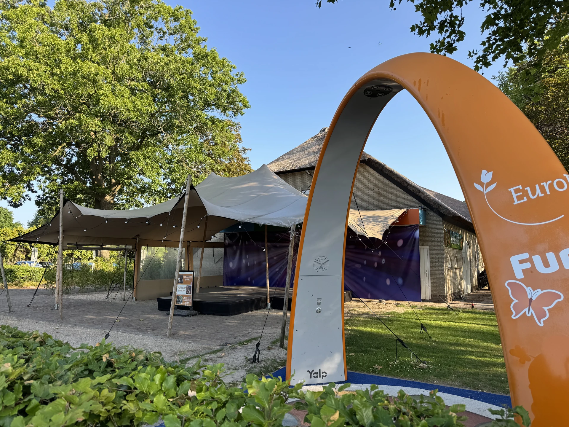 Reetgedecktes Zentralgebäude im EuroParcs Kaatsheuvel mit Stretchzelt, Bühne und orangefarbenem interaktivem Spielbogen davor