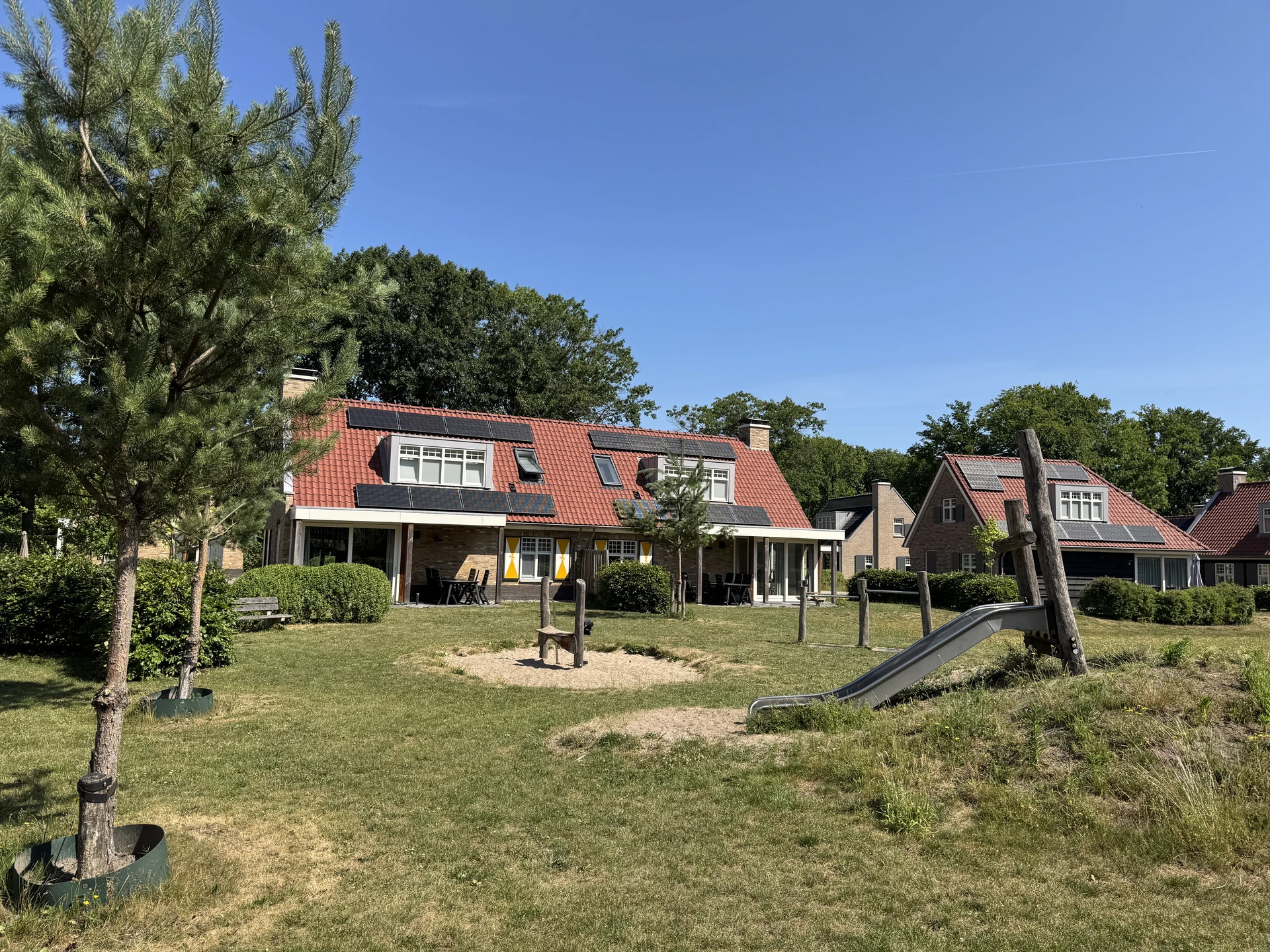 Spielplatz im Landal Kaatsheuvel mit Metallrutsche im Grashügel, Sandbereich und Federtier, dahinter Ferienhäuser