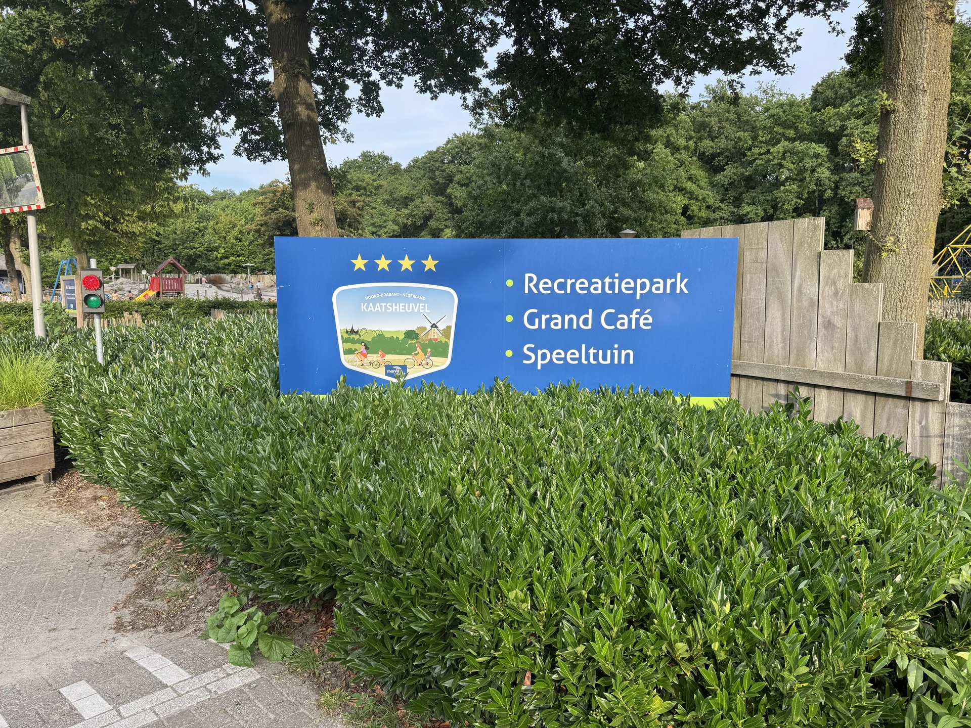 Eingangsschild von Marvilla Parks Kaatsheuvel mit den Angeboten Recreatiepark, Grand Café und Speeltuin, dahinter der Spielplatz mit Rutsche und Sandfläche