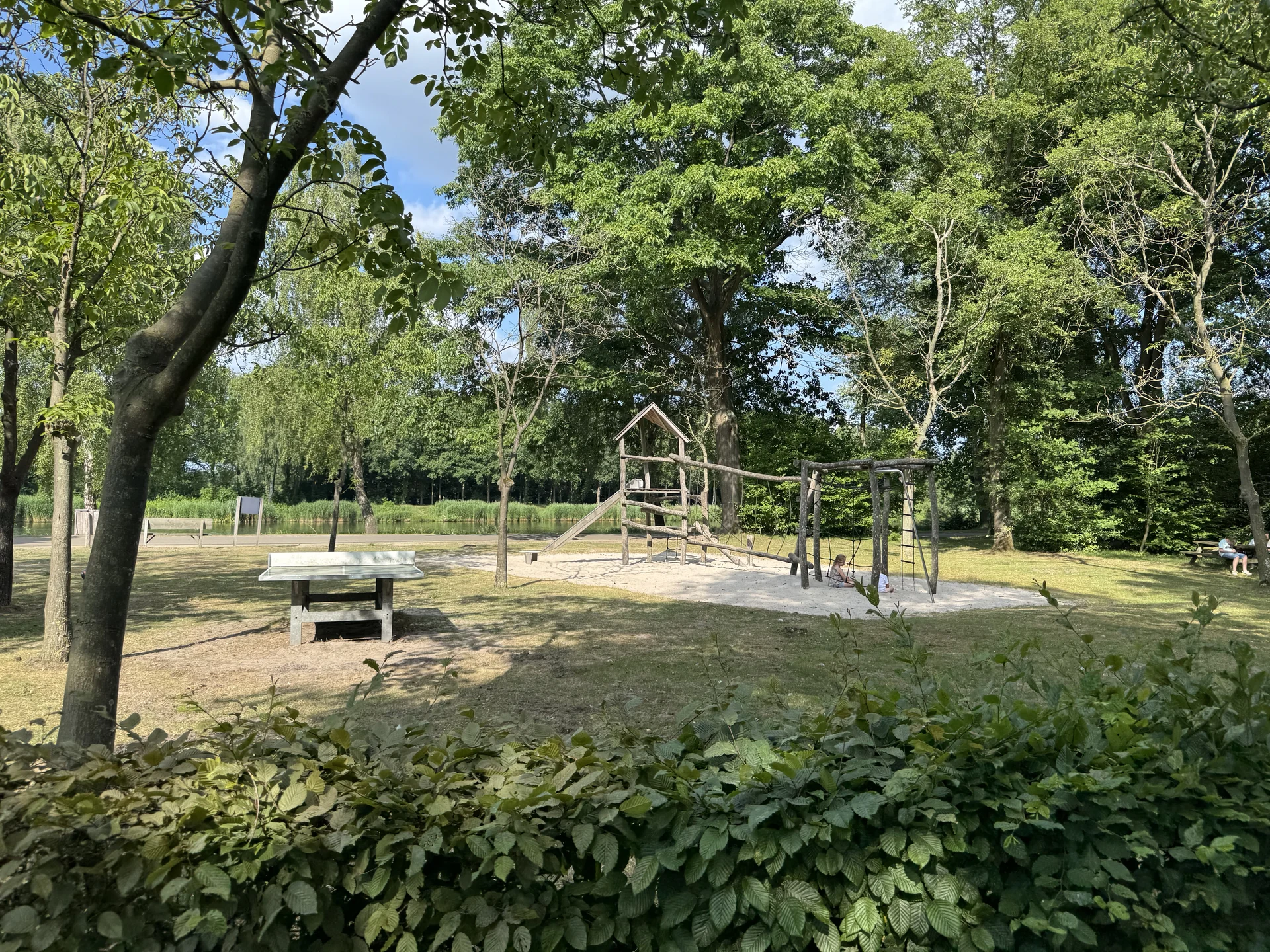 Naturholz-Spielplatz mit Rutsche und Klettergerüst auf Sand am Ufer im Lake Resort Beekse Bergen, daneben eine Tischtennisplatte