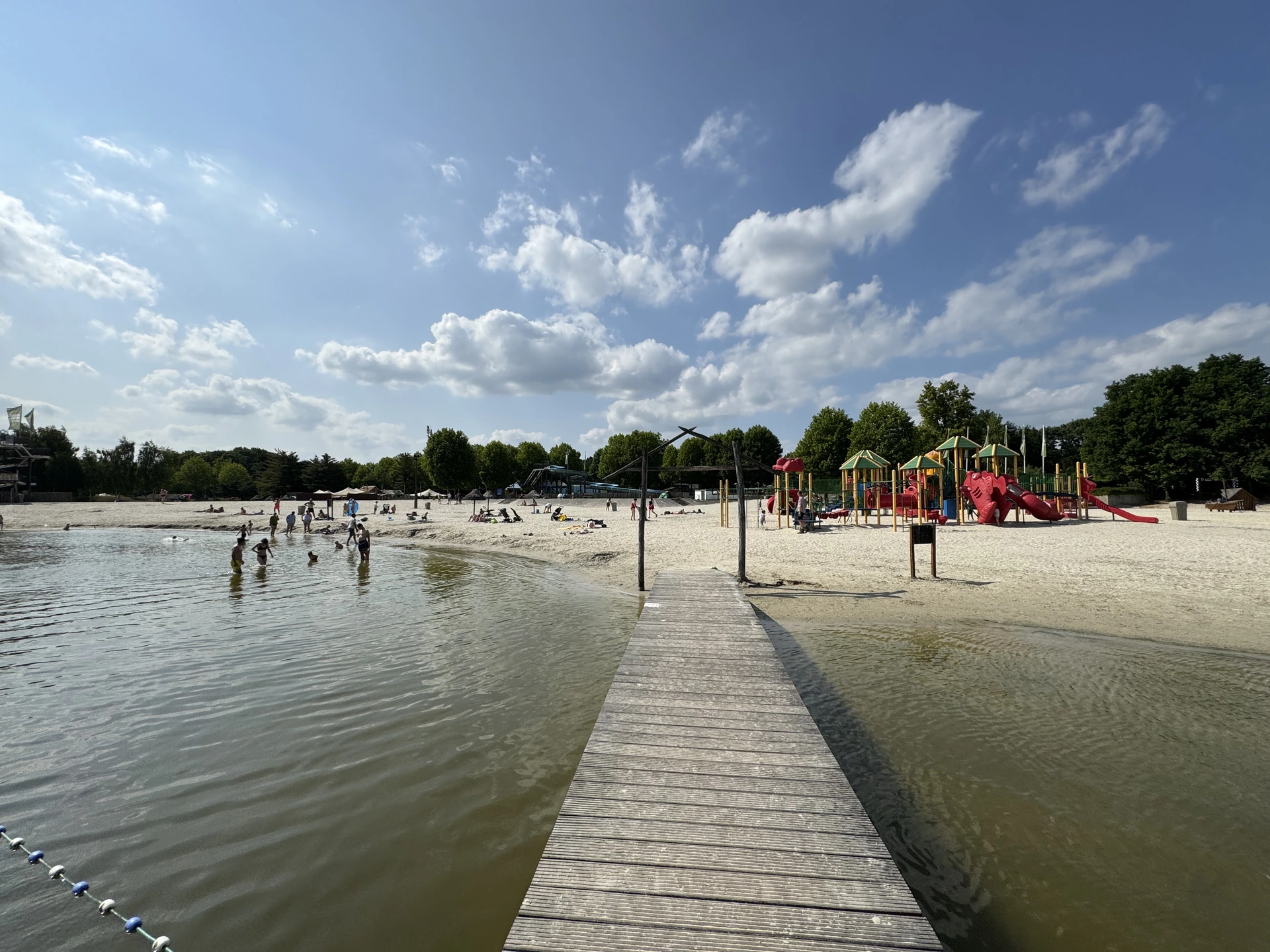 Blick vom Steg auf den Badestrand im Speelland Beekse Bergen mit Fahrgeschäften am Ufer